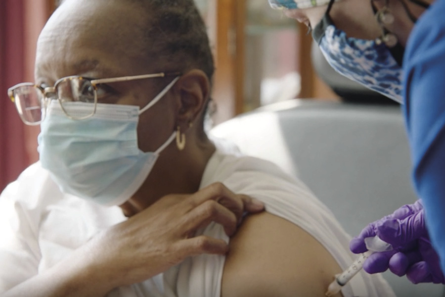 An older Black woman wearing glasses and a mask lifts the sleeve of her shirt to expose her upper arm as a home care provider injects COVID-19 vaccine.